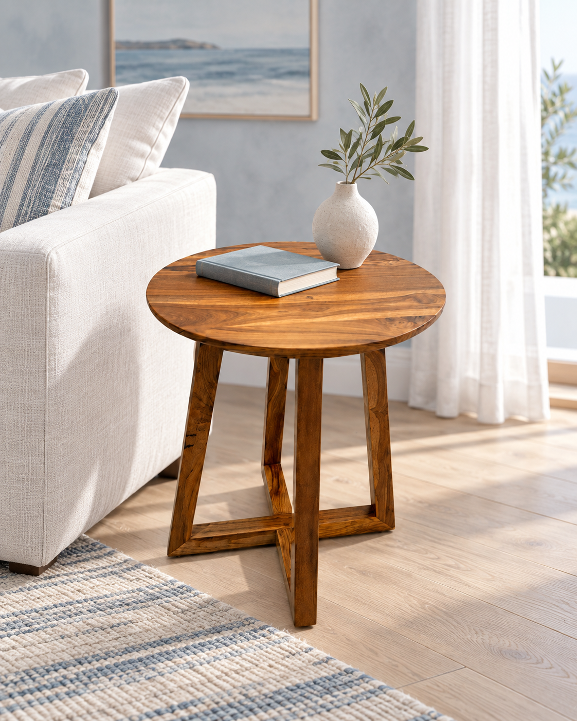 A round wooden end table with a warm medium-brown finish, styled in a bright living room next to a white sofa. The table holds a gray book and a small white vase with greenery.