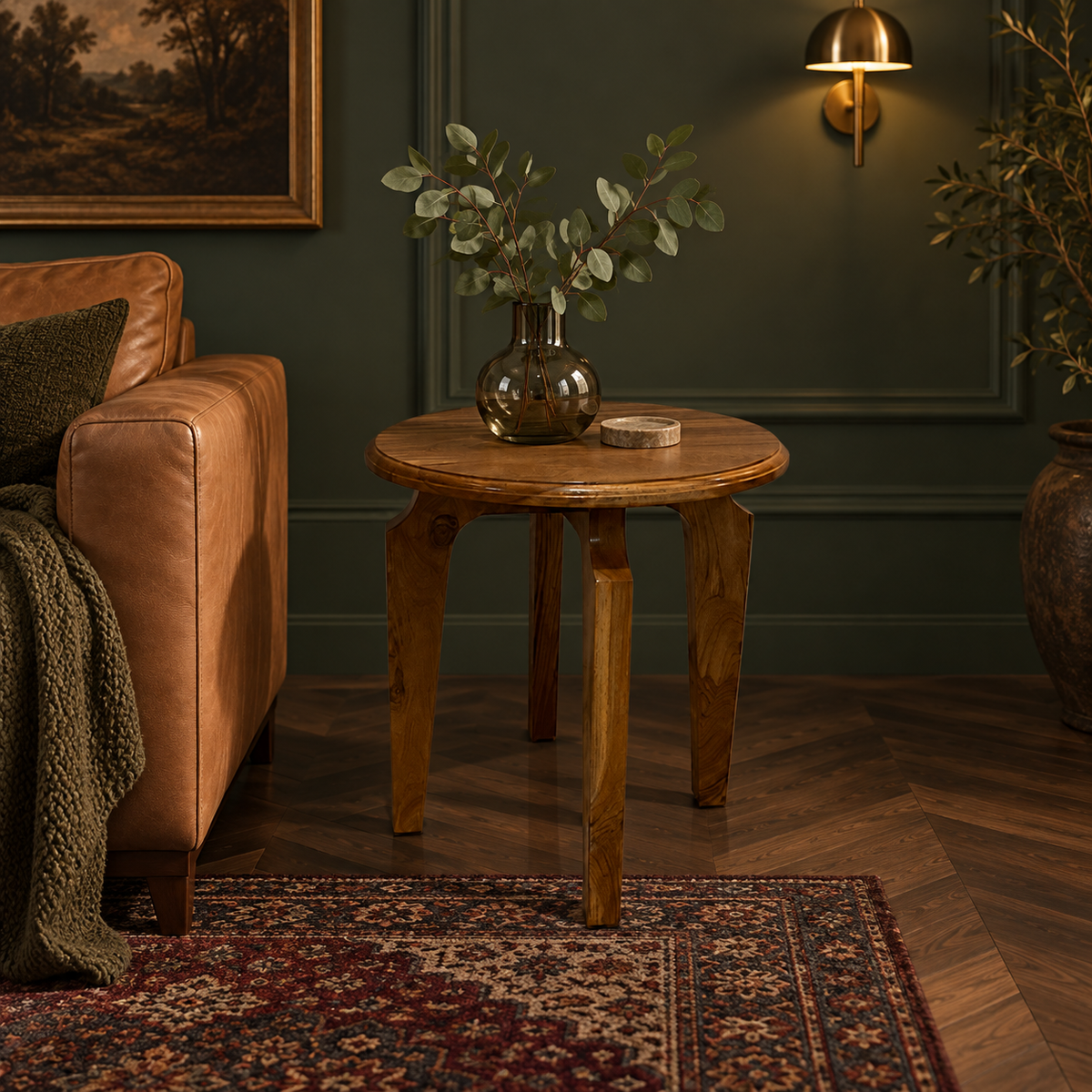 A round wooden side table with three legs in a cozy living room setting, styled with a glass vase of eucalyptus and a small coaster, next to a brown leather sofa.