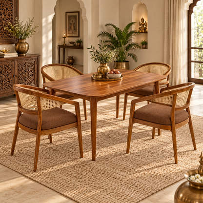 A wooden dining table with four matching armchairs featuring woven cane backs and brown upholstered seats, styled in a warm, sunlit traditional interior with a woven rug and decorative plants.